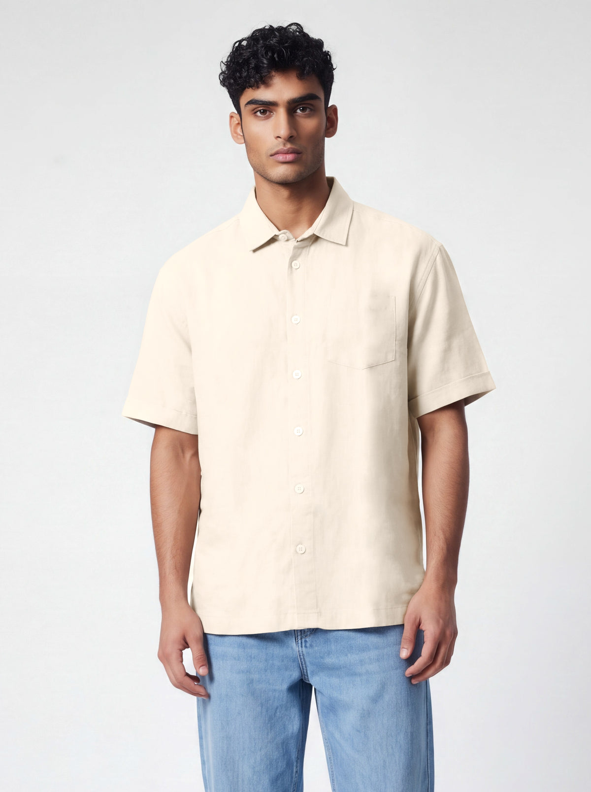 A man wearing a light beige short-sleeve button-up shirt and blue jeans, standing against a light gray studio background.
