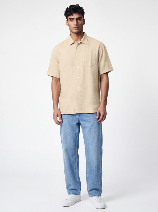 A man modeling a light beige short-sleeve linen button-up shirt with light blue jeans and white sneakers, standing against a plain white studio background.