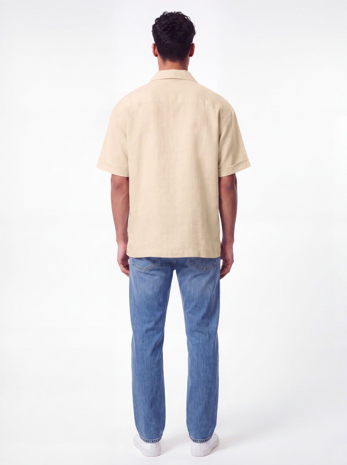 A man viewed from behind wearing a light beige short-sleeve button-up shirt and blue jeans against a plain white studio background.