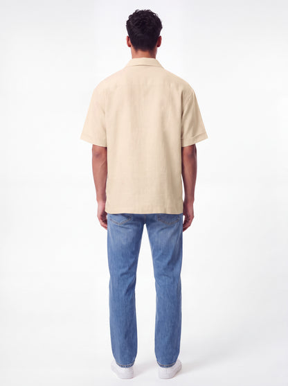 A man viewed from behind wearing a light beige short-sleeve button-up shirt and blue jeans against a plain white studio background.