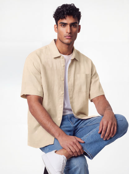 A man wearing a beige short-sleeve button-up shirt over a white t-shirt, paired with blue jeans and white sneakers, posing against a light gray studio background.