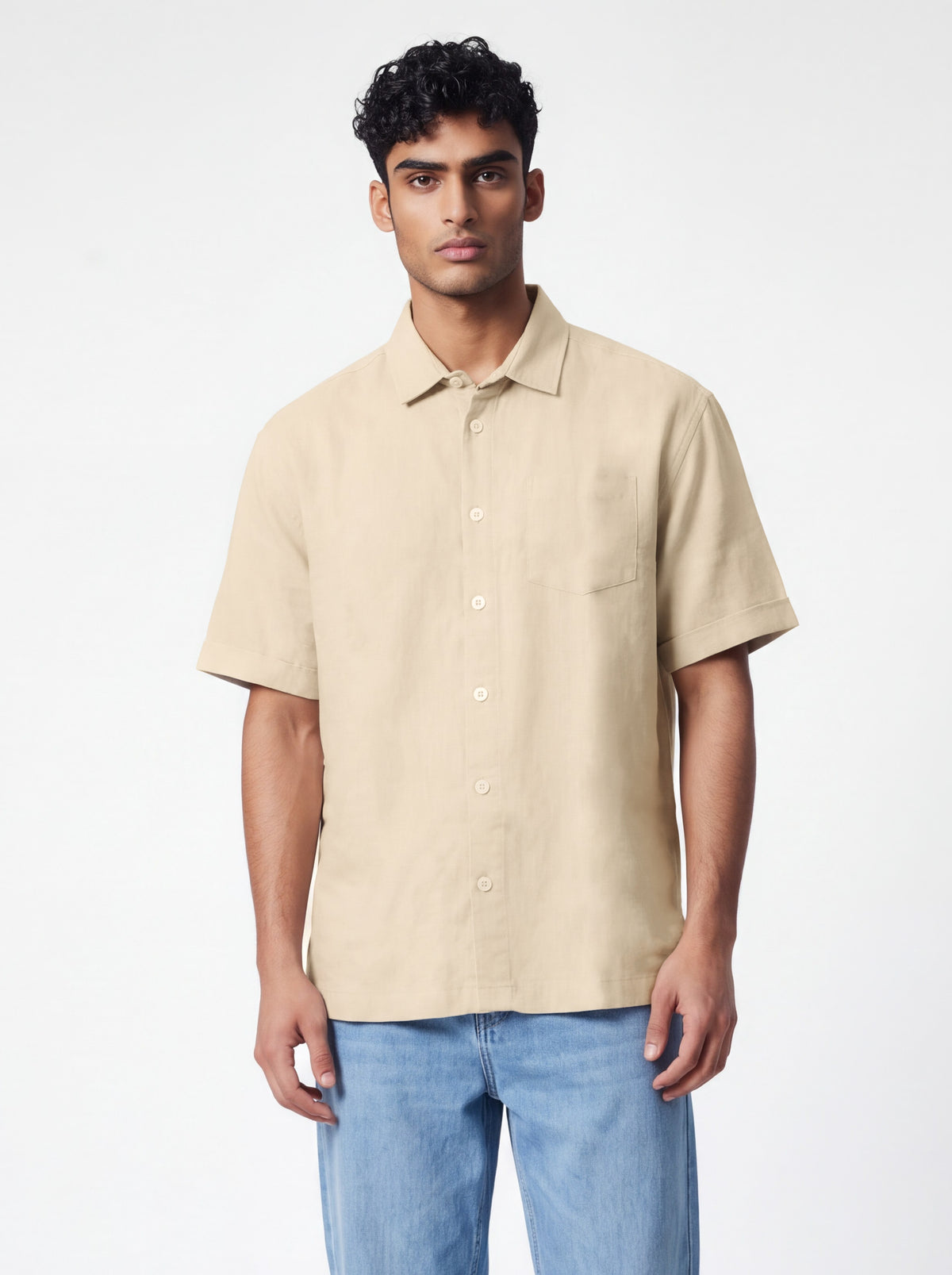 A man wearing a beige short-sleeve button-up shirt and light blue jeans, standing against a plain white studio background.