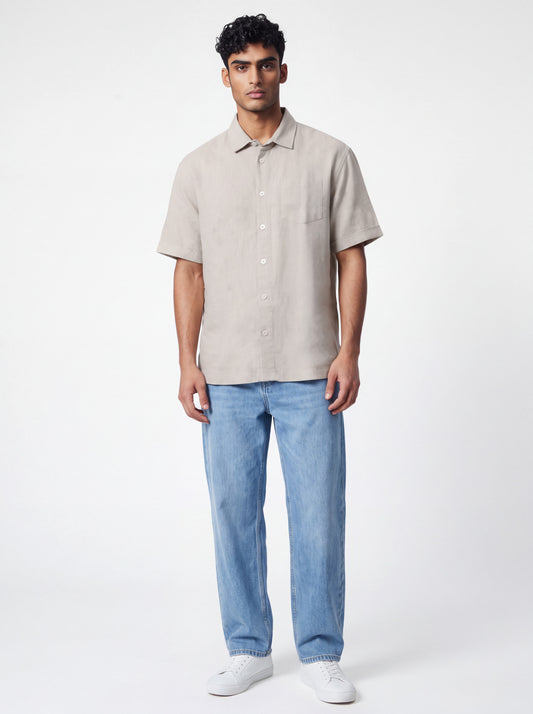 A man modeling a light beige short-sleeve linen button-up shirt with light blue jeans and white sneakers, standing against a plain white studio background.
