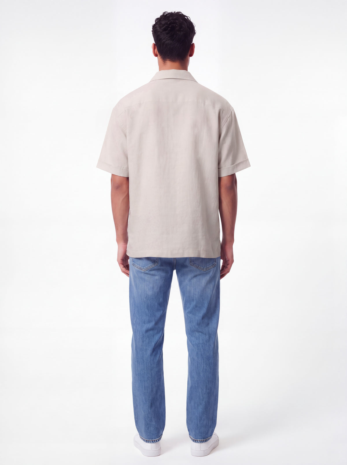 A man viewed from behind wearing a light beige short-sleeve button-up shirt and blue jeans against a plain white studio background.