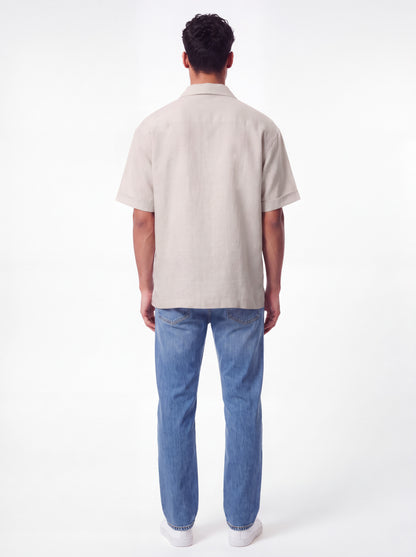 A man viewed from behind wearing a light beige short-sleeve button-up shirt and blue jeans against a plain white studio background.
