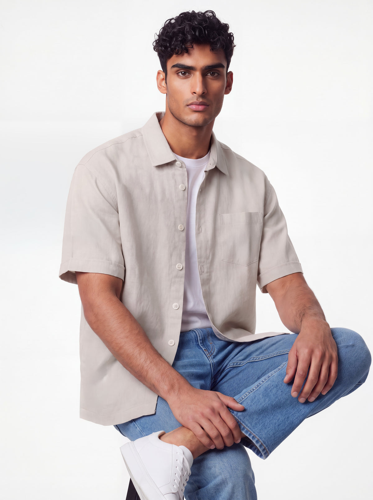 A man wearing a light beige short-sleeve button-up shirt over a white t-shirt, paired with blue jeans and white sneakers, posing against a clean white studio background.
