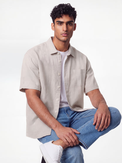 A man wearing a light beige short-sleeve button-up shirt over a white t-shirt, paired with blue jeans and white sneakers, posing against a clean white studio background.