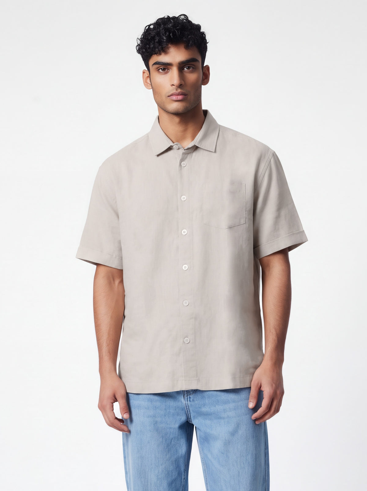 A man wearing a light beige short-sleeve button-up shirt and blue jeans, standing against a plain white studio background.