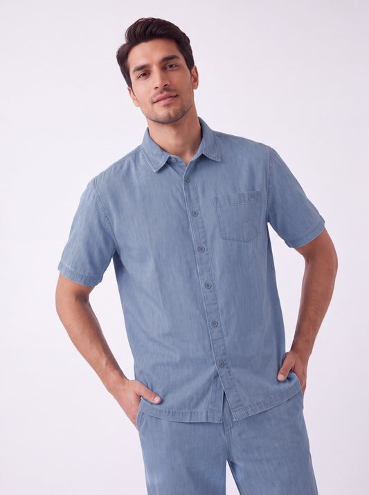 A man wearing a light blue short-sleeve button-up shirt and matching light blue pants, standing against a plain white studio background with his hands in his pockets.