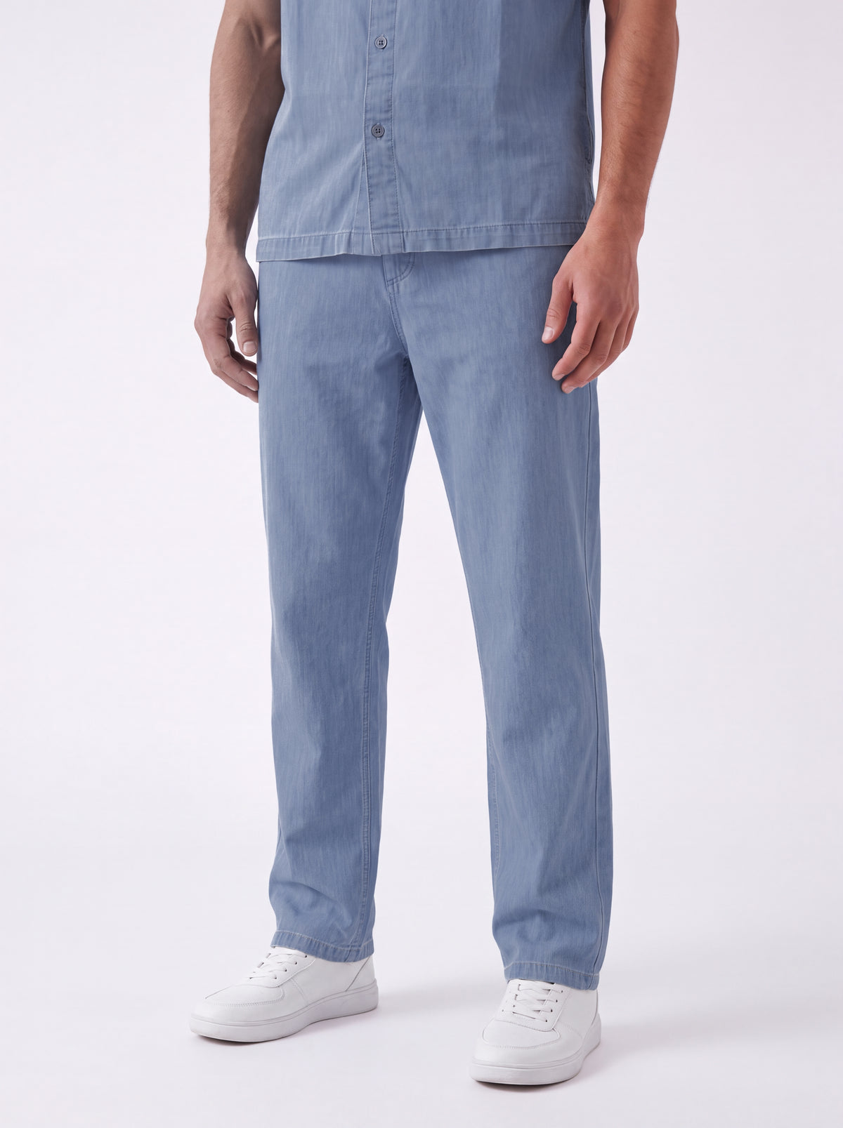A model wearing a light blue denim shirt and matching light blue denim trousers, paired with white sneakers, shown from the chest down against a plain white studio background.
