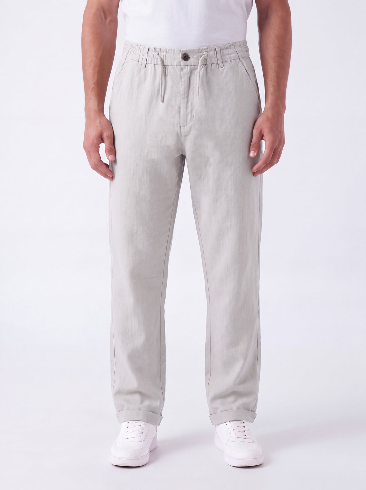 A man wearing light gray drawstring trousers, a white t-shirt, and white sneakers, standing against a plain white studio background.
