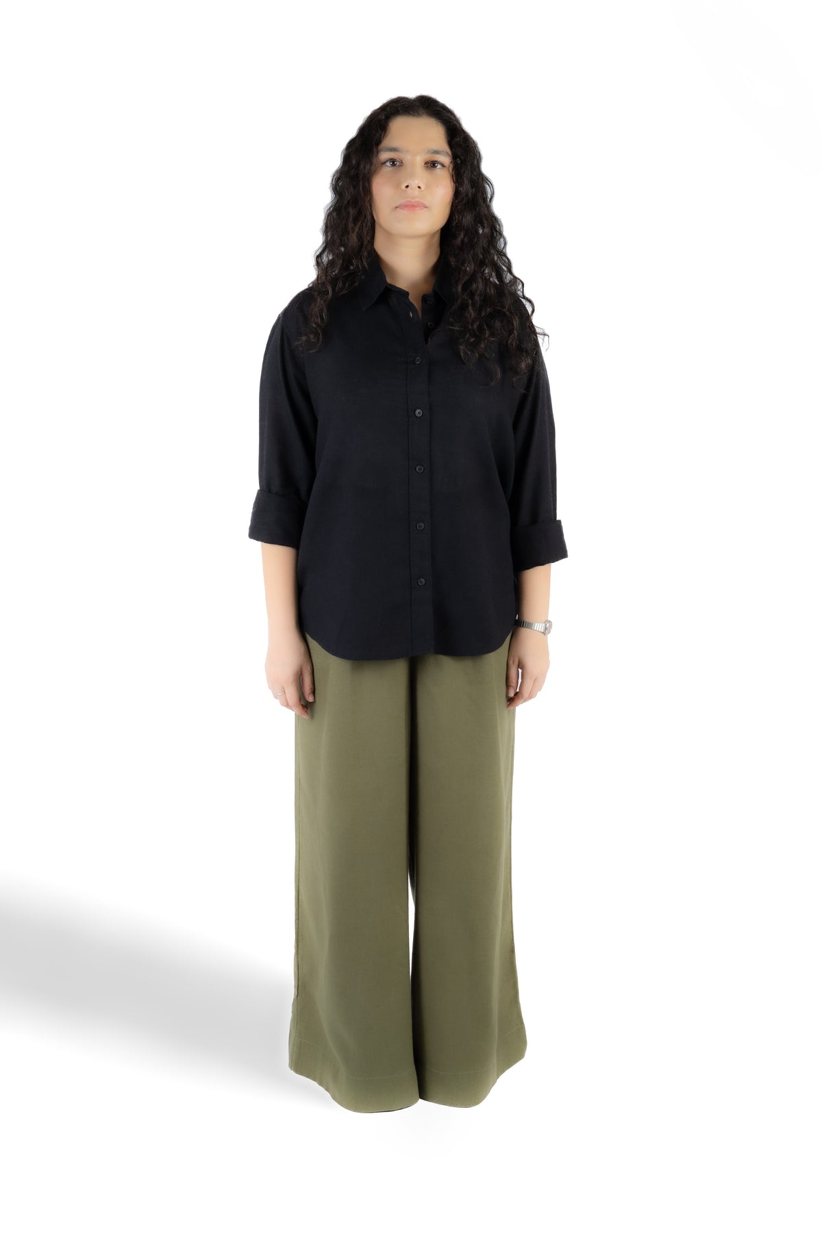 A woman stands against a white studio background wearing a black button-front shirt with rolled sleeves and olive green wide-leg pants.