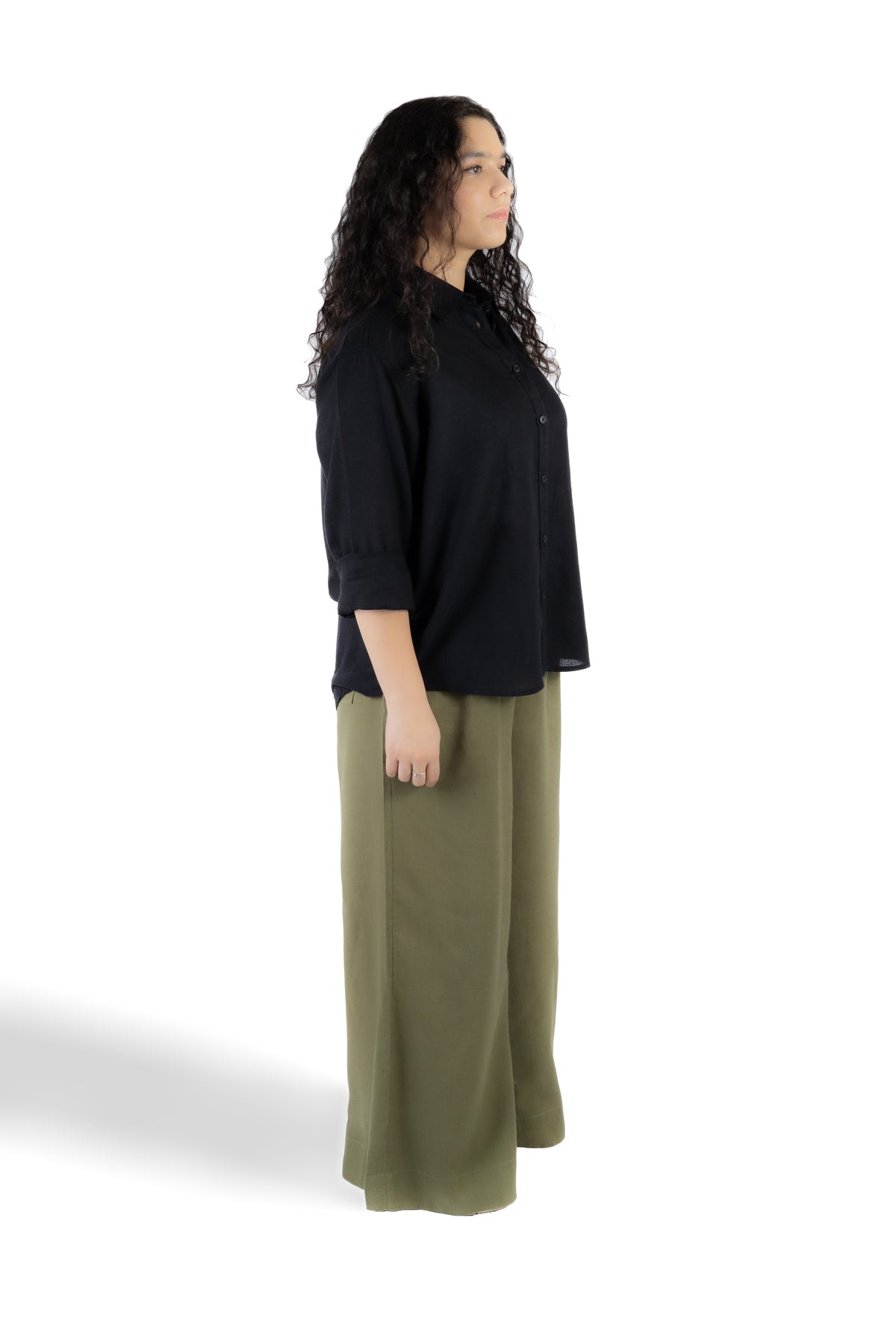 A woman shown in side profile wearing a black three-quarter sleeve button-front top and olive green wide-leg pants against a plain white studio background.