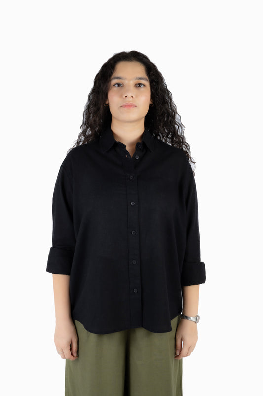 A woman with long curly dark hair wearing a black button-down shirt with rolled-up sleeves and olive green pants, standing against a plain white background.