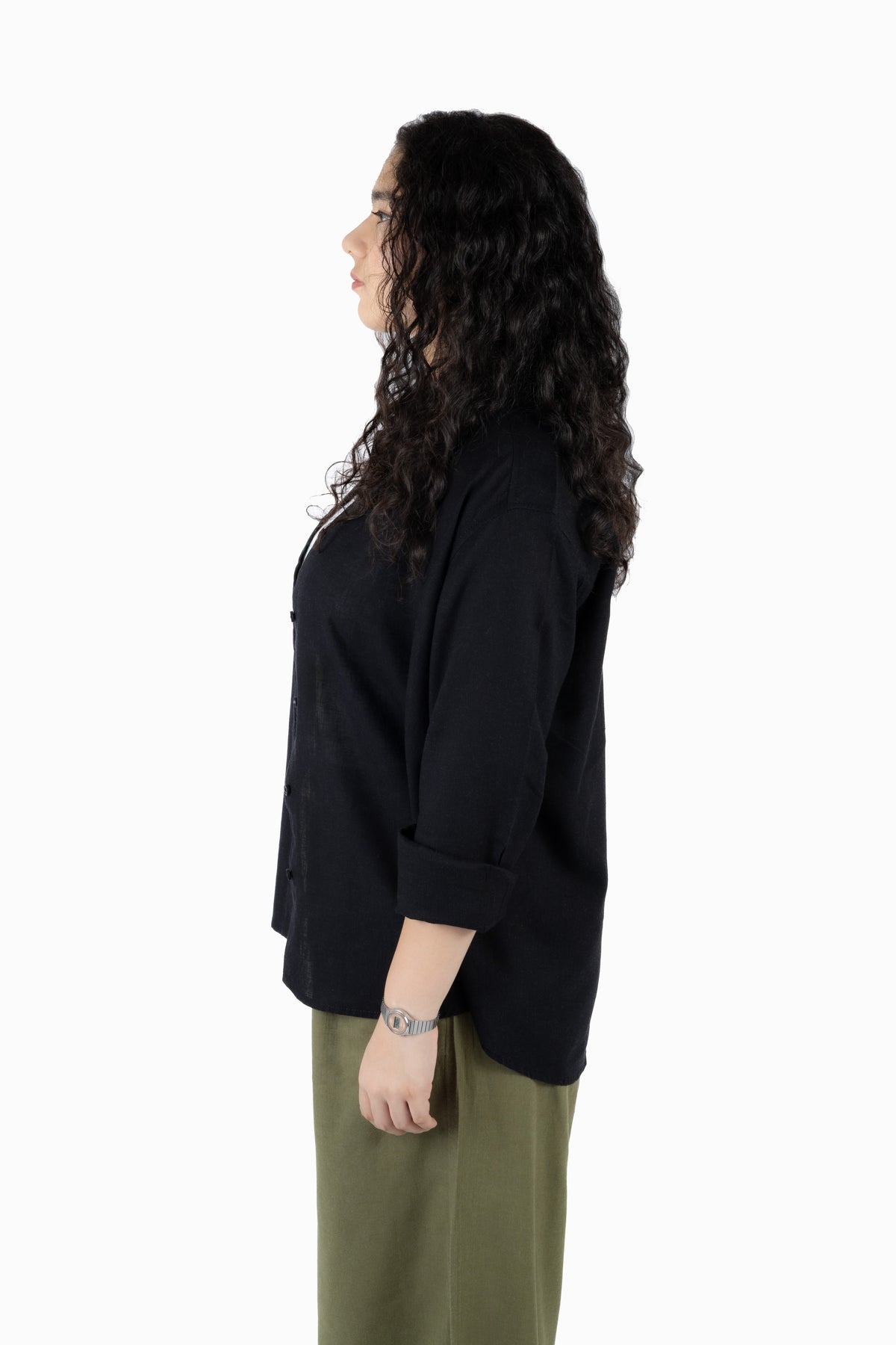 A woman with long, curly dark hair is shown in profile wearing a black button-up shirt with rolled-up sleeves and olive green pants, standing against a solid white background.