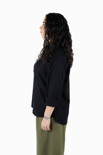 A woman with long, curly dark hair is shown in profile wearing a black button-up shirt with rolled-up sleeves and olive green pants, standing against a solid white background.