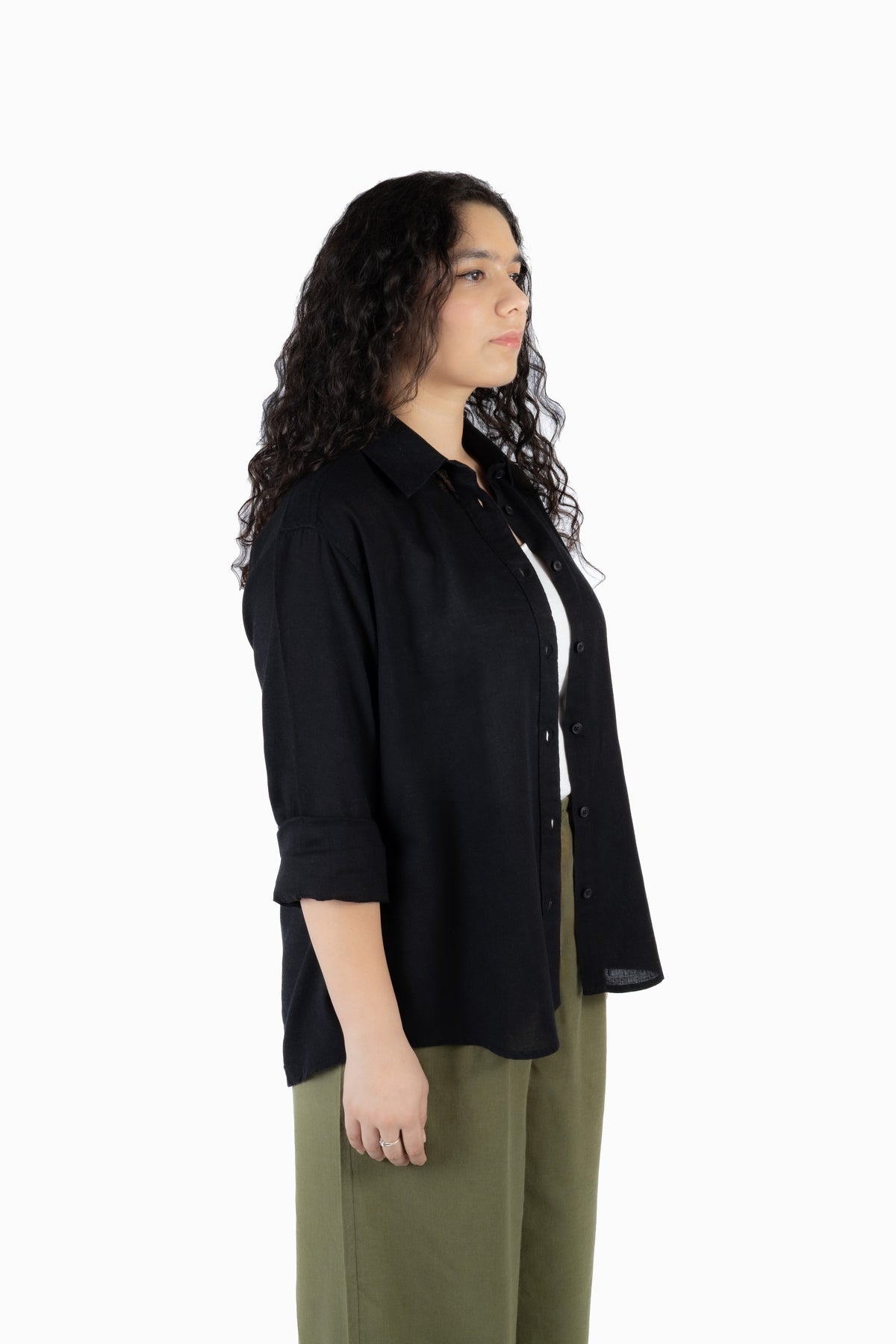 A woman with long, curly dark hair is shown in profile wearing a black button-up shirt over a white top and olive green pants, photographed against a solid white studio background.