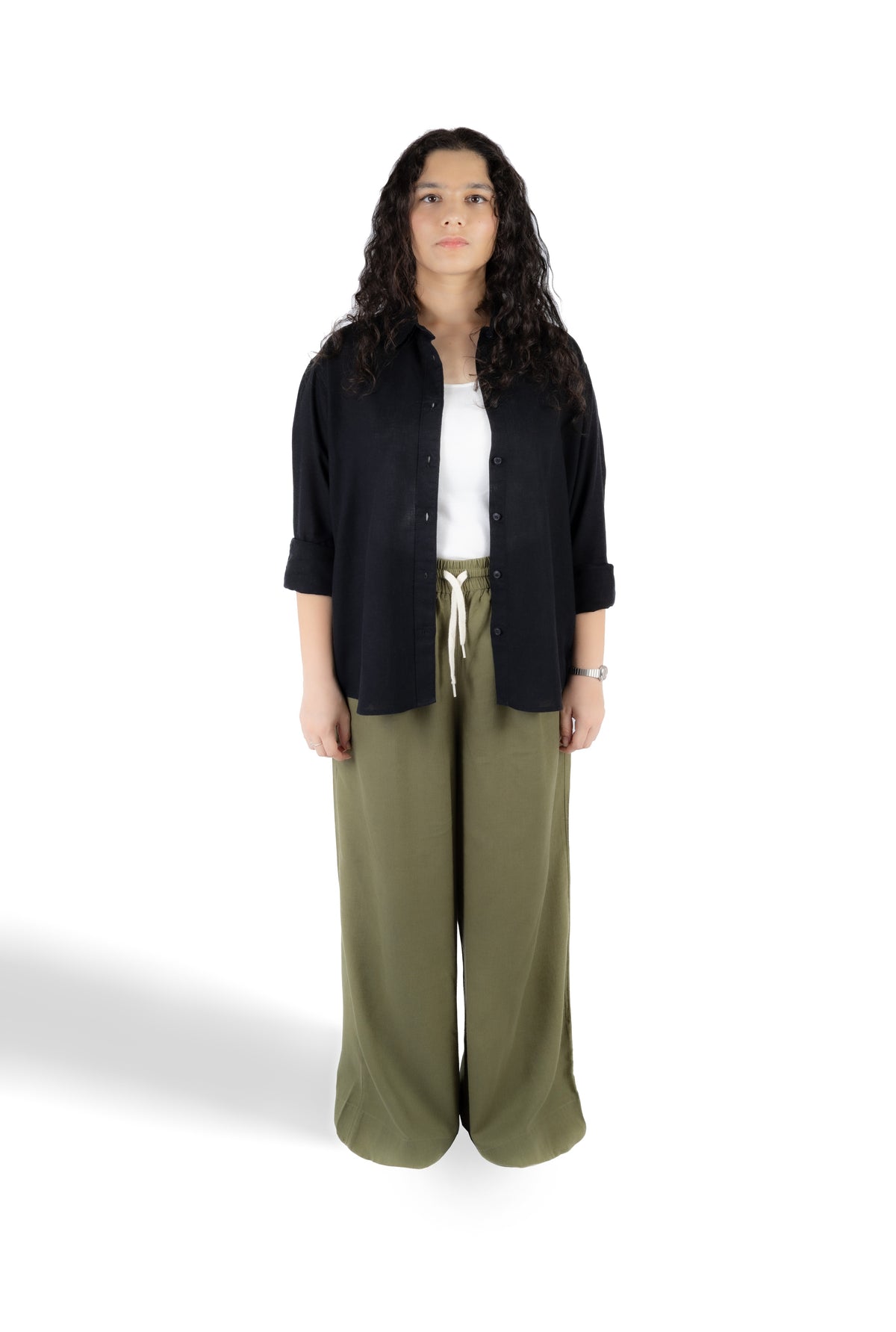 A woman stands against a white studio background wearing a black button-up shirt over a white top, paired with olive green wide-leg pants.