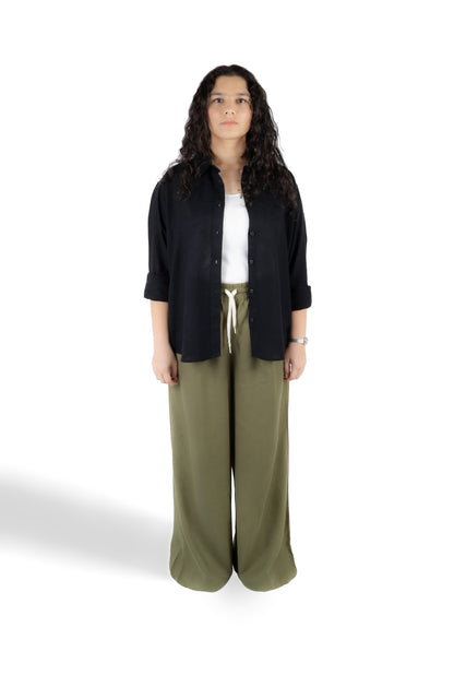 A woman stands against a white studio background wearing a black button-up shirt over a white top, paired with olive green wide-leg pants.