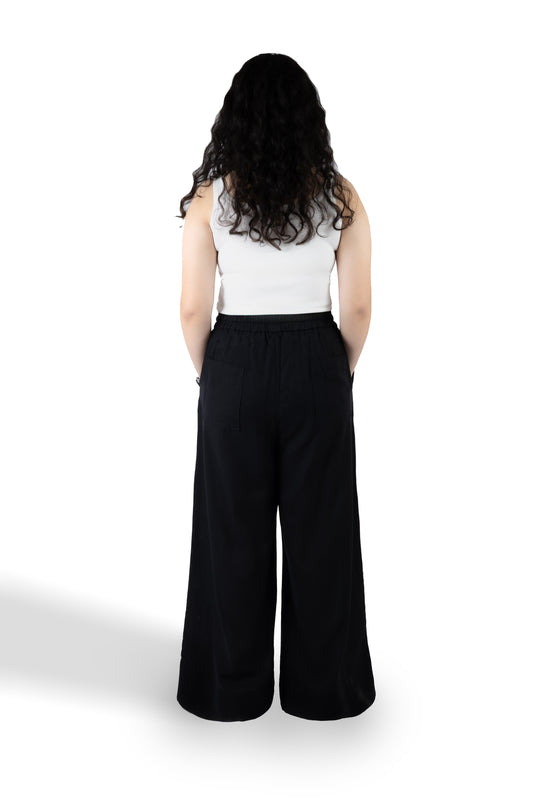 A woman with long, curly black hair is shown from behind wearing a white sleeveless top and black wide-leg pants, standing against a plain white background.