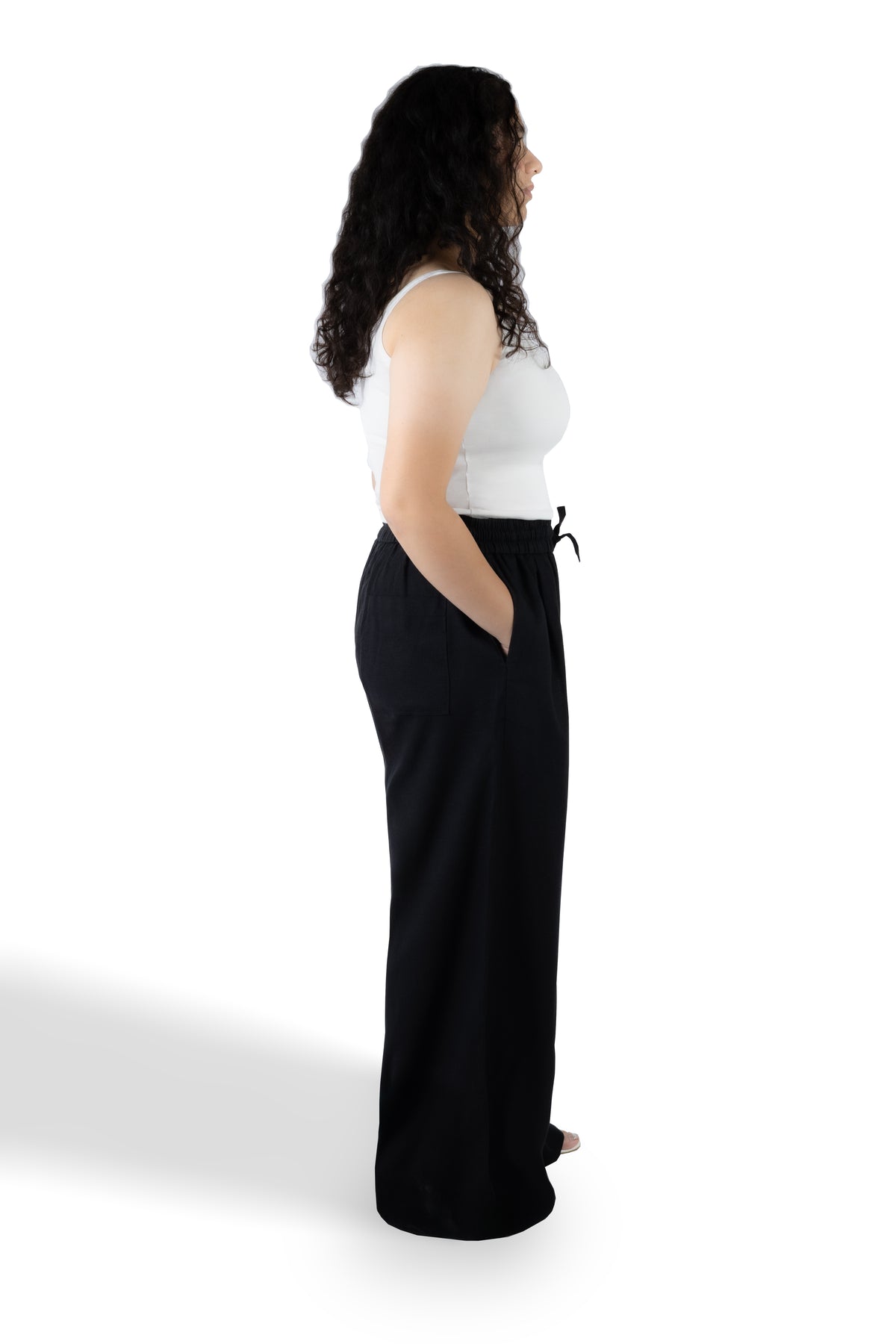 A woman shown from the side profile wearing a white sleeveless top and black wide-leg pants with side pockets, standing against a plain white studio background.
