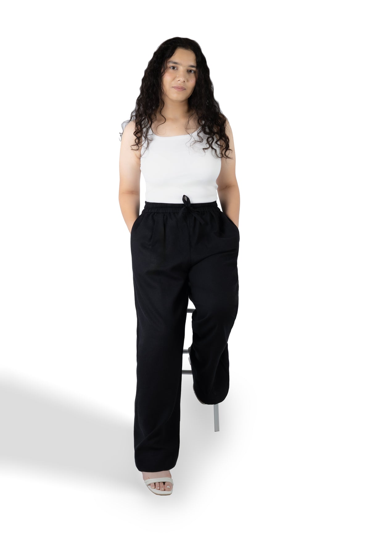 A woman stands on a white studio background wearing a white sleeveless top and loose black drawstring pants, with one leg bent over on a small stool.