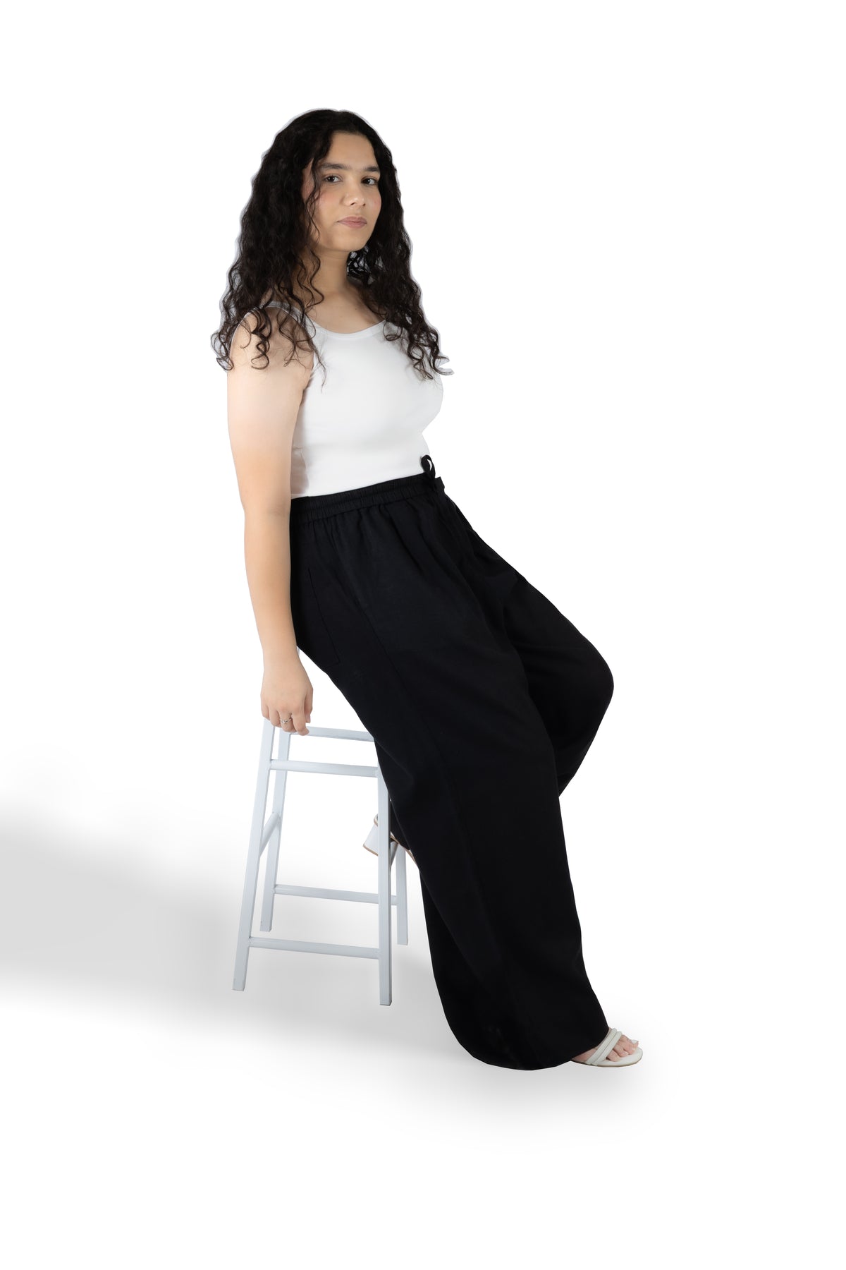 A woman with long curly dark hair sits on a white stool against a white studio background, wearing a white sleeveless top and loose black wide-leg pants.