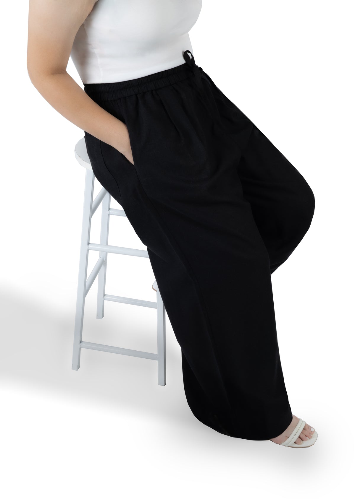 A person wearing black wide-leg pants with a white top, sitting on a white stool against a white background.