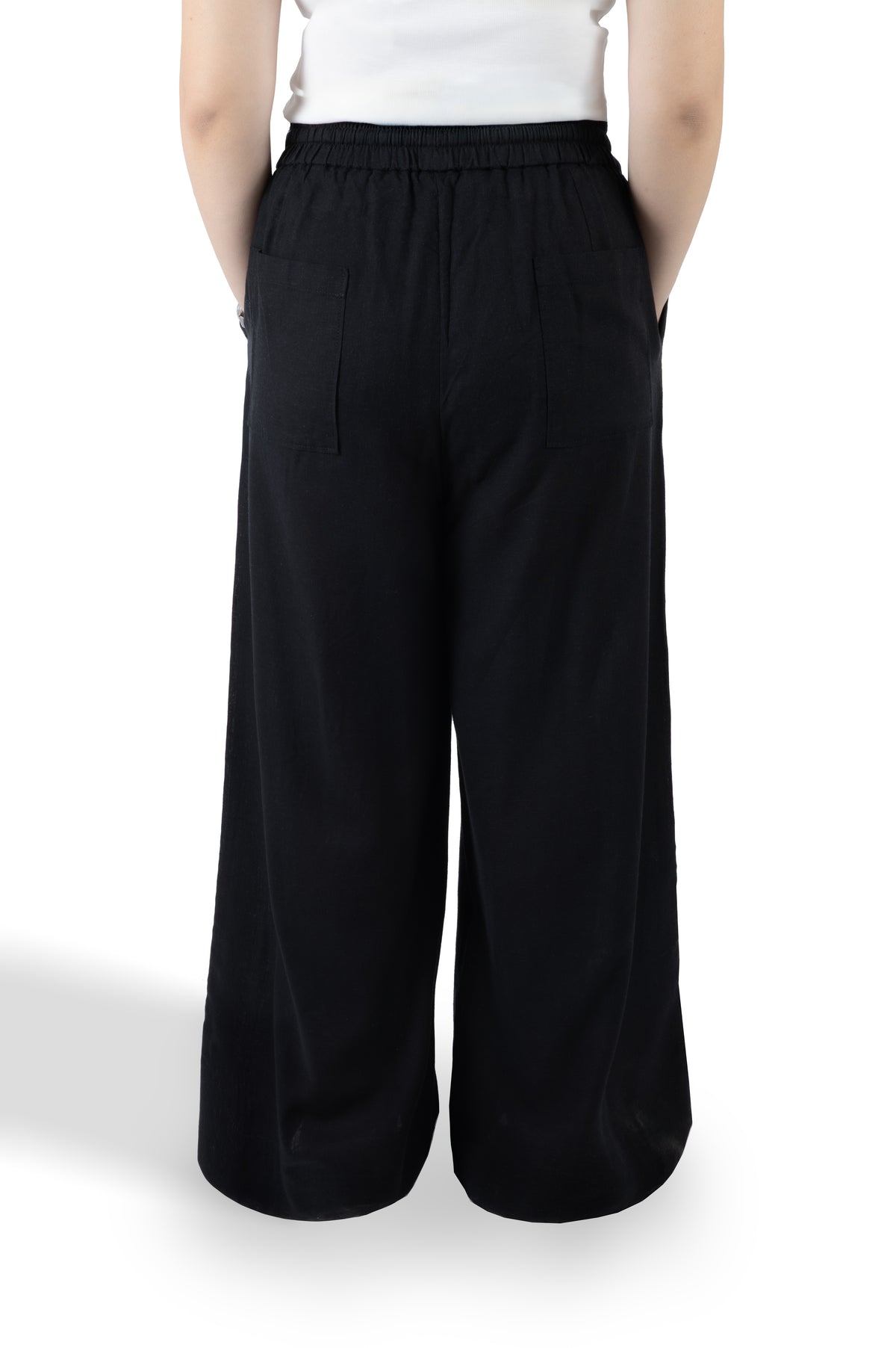 A person shown from the back wearing loose black wide-leg pants with an elastic waistband and side pockets, paired with a white top, against a plain white studio background.