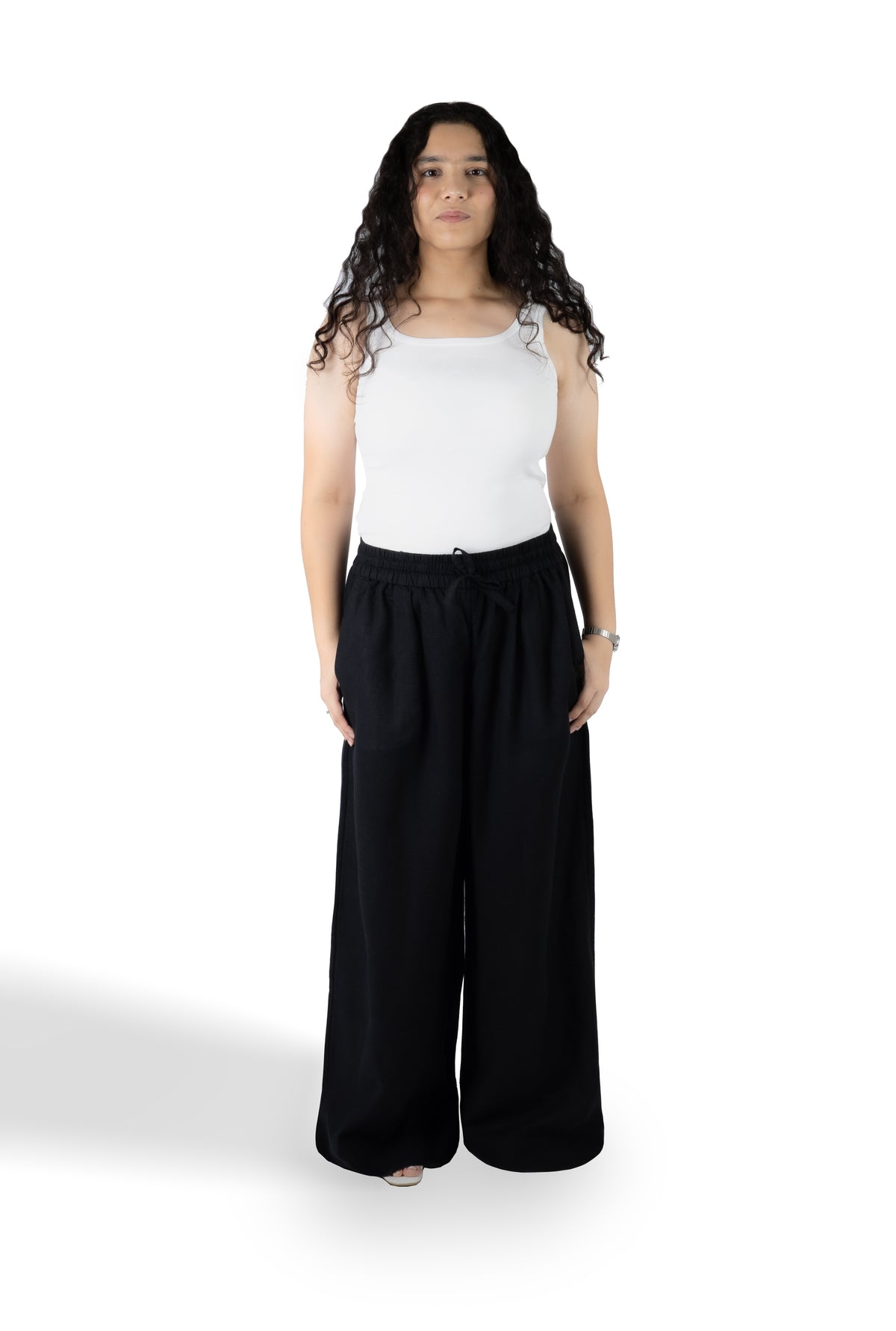 A woman stands against a white studio background wearing a white sleeveless top and loose black wide-leg pants.