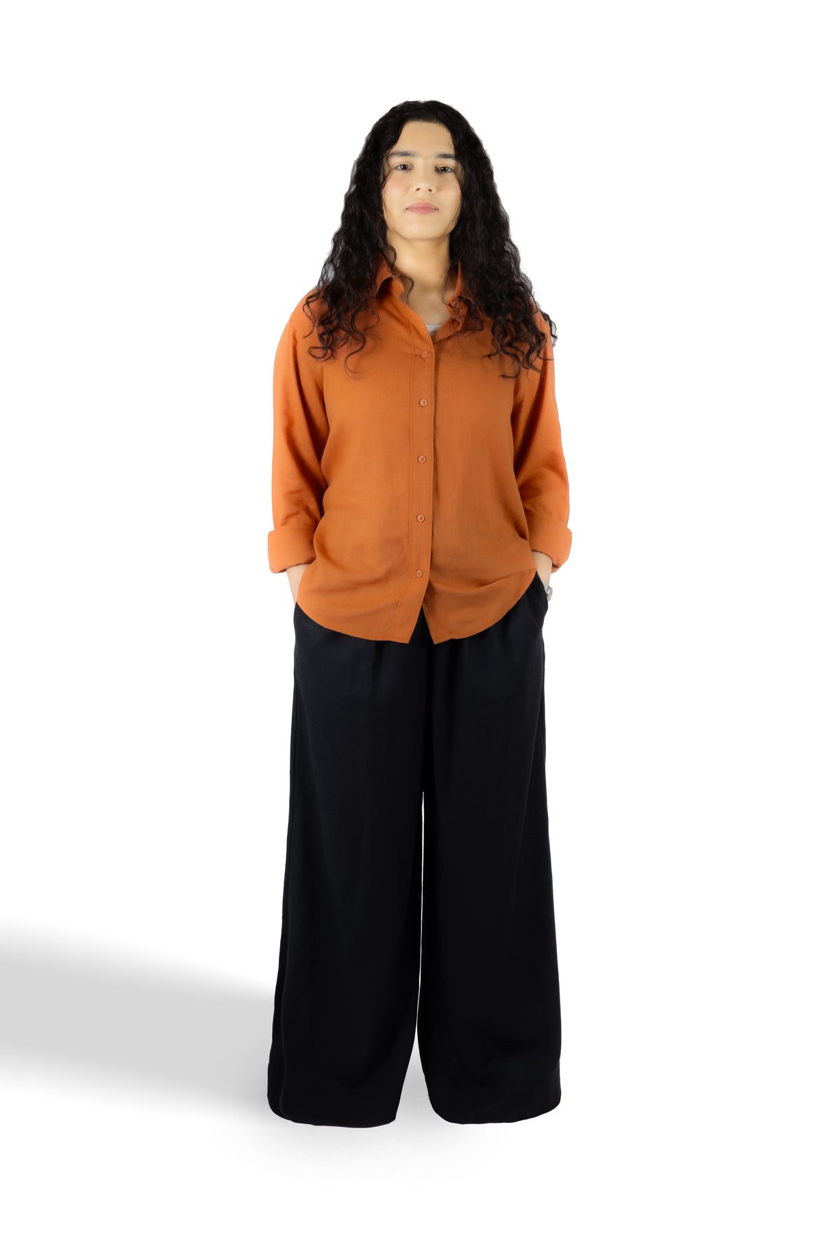 A woman modeling a long-sleeve button-up shirt in burnt orange paired with wide-leg black pants, standing against a plain white studio background.