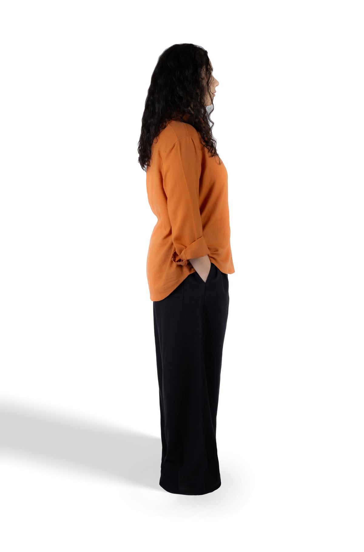 A woman shown in profile wearing a long-sleeve burnt orange top and black wide-leg pants, standing against a plain white studio background.