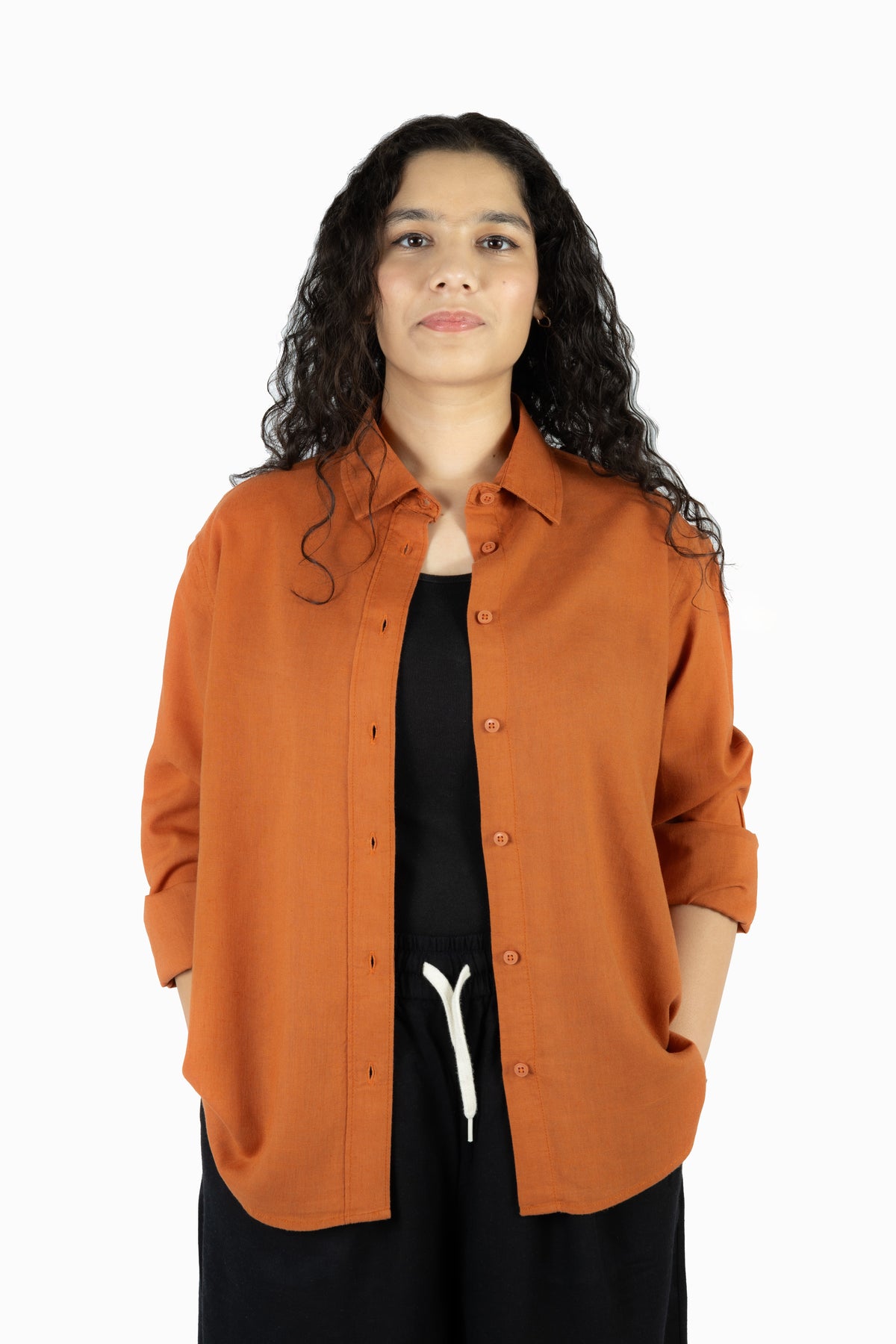 A woman with long, curly dark hair wearing an orange-brown button-up shirt over a black top and black pants, standing against a plain white background.