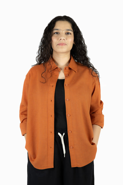 A woman with long, curly dark hair wearing an orange-brown button-up shirt over a black top and black pants, standing against a plain white background.