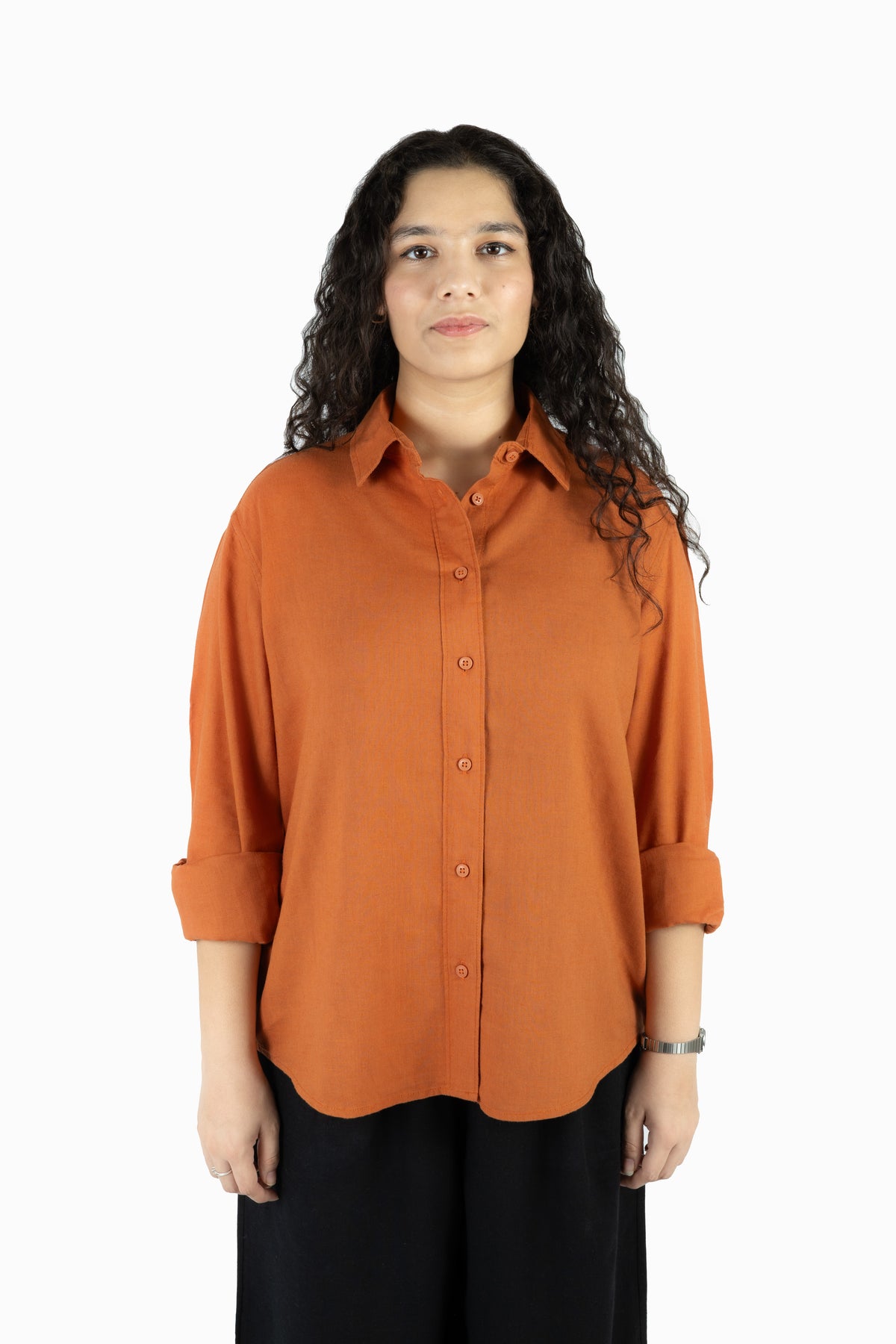 A woman with long, curly dark hair wearing a rust-orange button-down shirt with rolled-up sleeves and black pants, standing against a plain white background.