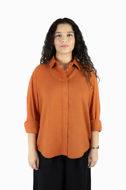A woman with long, curly dark hair wearing a rust-orange button-down shirt with rolled-up sleeves and black pants, standing against a plain white background.