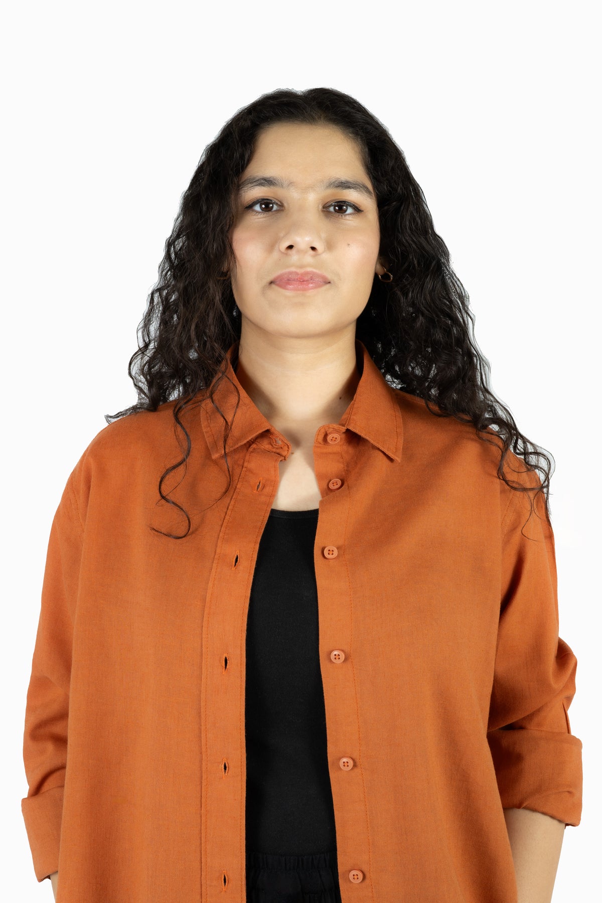 A woman with long, curly dark hair wearing a rust-orange button-up shirt over a black top, photographed against a solid white background.
