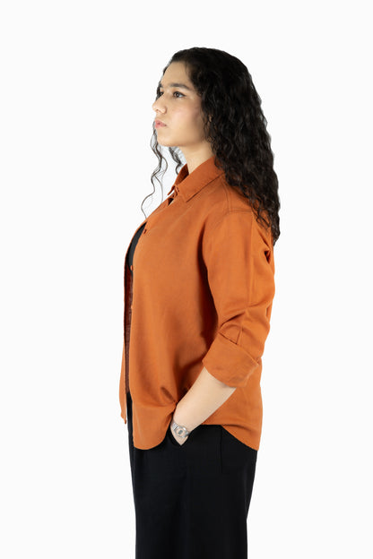 A woman with long, curly dark hair is shown in profile wearing a burnt orange button-up shirt and black pants against a plain white background.