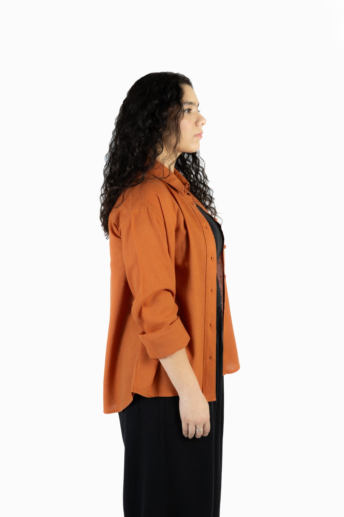 A woman with long, curly dark hair is shown in profile wearing a rust-orange button-up shirt over a black top and black pants, photographed against a solid white background.