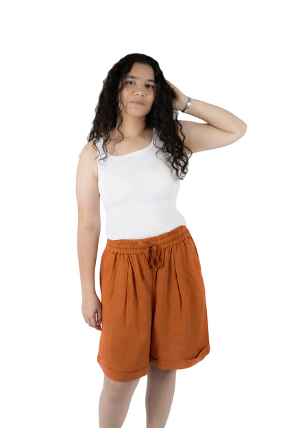 A woman modeling a pair of rust-orange drawstring shorts and a white tank top against a solid white background.