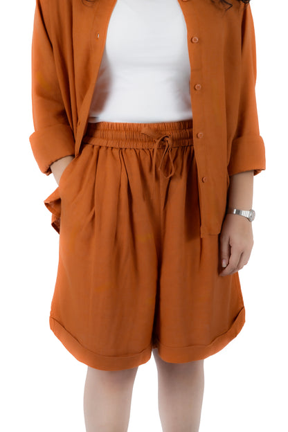 A person wearing a rust-orange button-front overshirt and matching high-waisted drawstring shorts over a white top, shown from the shoulders to mid-thigh on a plain white background.