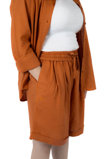 A model wearing a rust-orange linen-blend set consisting of a button-up shirt and matching drawstring shorts, styled over a white top against a plain white background.