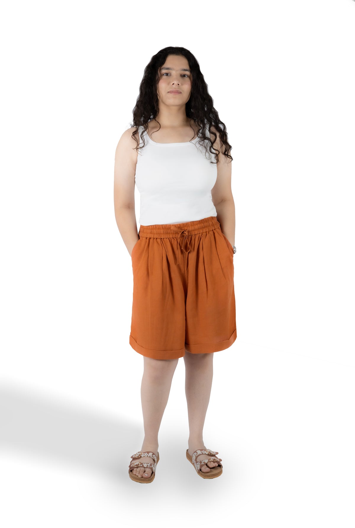A woman modeling a pair of rust-orange drawstring shorts with a white tank top and brown sandals, standing against a plain white studio background.