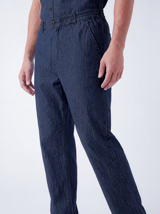 A close-up of a person wearing dark blue denim overalls or jumpsuit pants, showing the waist, front pockets, and upper legs against a clean light gray studio background.