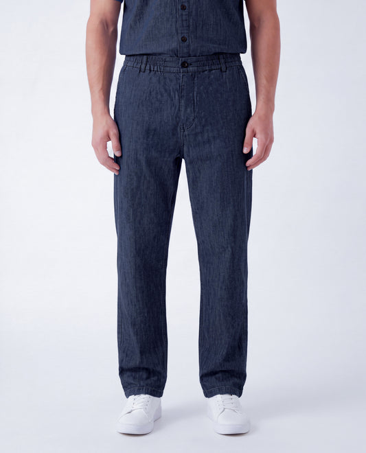 A person wearing dark indigo denim pants and a matching dark denim shirt, paired with white sneakers, photographed against a clean light gray studio background.