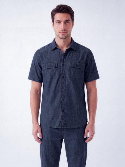 A man wearing a dark indigo short-sleeve button-up shirt with two chest pockets and matching dark jeans, standing against a light gray studio background.