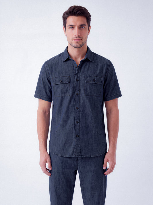 A man wearing a dark indigo short-sleeve button-up shirt with two chest pockets and matching dark jeans, standing against a light gray studio background.