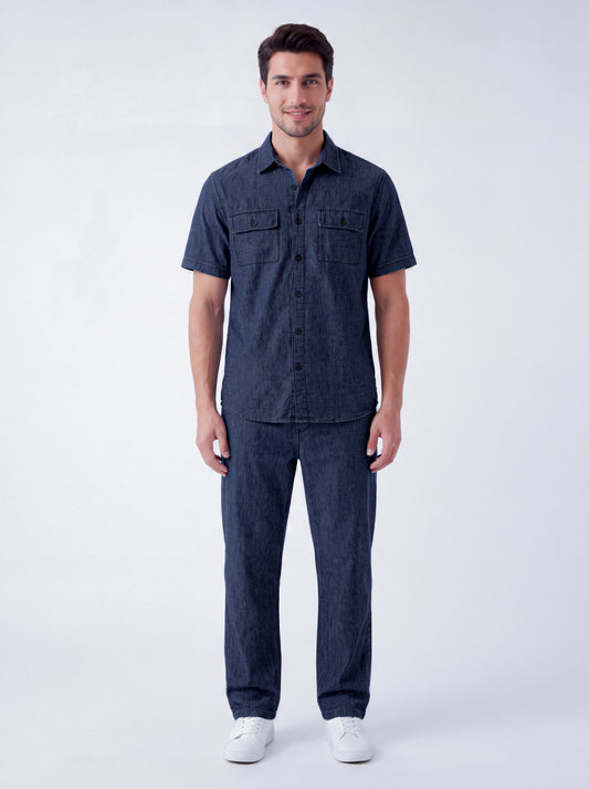 A man modeling a dark navy blue short-sleeve button-up shirt and matching navy blue jeans, standing against a light gray studio background.