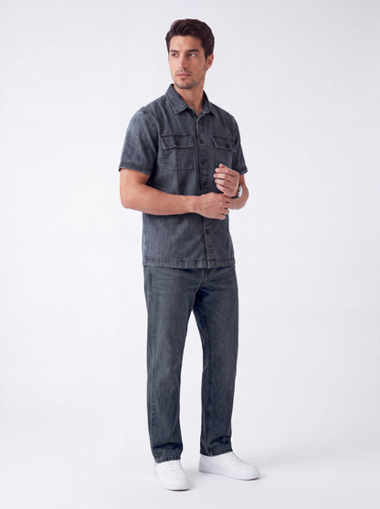 A man modeling a dark gray short-sleeve denim shirt and matching dark gray jeans with white sneakers, standing against a light gray studio background.