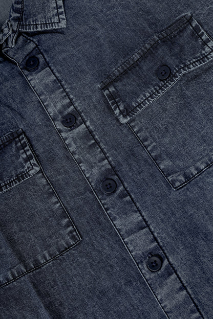 Close-up of a dark washed denim button-up shirt showing the chest pocket, button closure, and fabric texture.